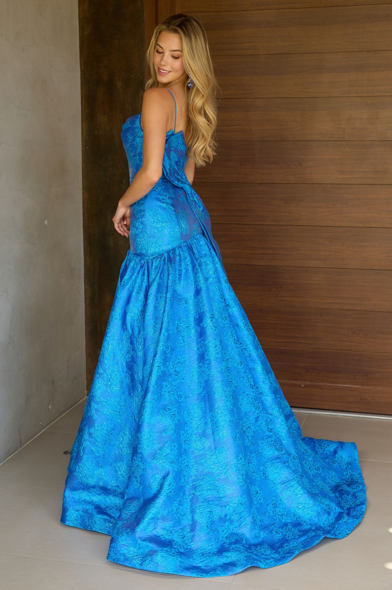 A woman stands indoors, looking over her shoulder in the Ava Presley 26419 Drop-Waist Long Print Prom Gown.